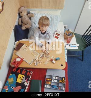 Un garçon au lit dans le 1960s. Il a probablement un froid et a besoin de rester à la maison. Cependant, il n'y a pas de grand besoin pour lui. Il garde ses jouets à portée de main comme les voitures, les bateaux et les animaux en peluche. Dans l'image il met des puzzles ensemble. Suède 1969 Banque D'Images