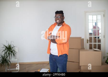 Homme noir souriant posant dans la chambre avec des boîtes en carton le jour du déménagement Banque D'Images