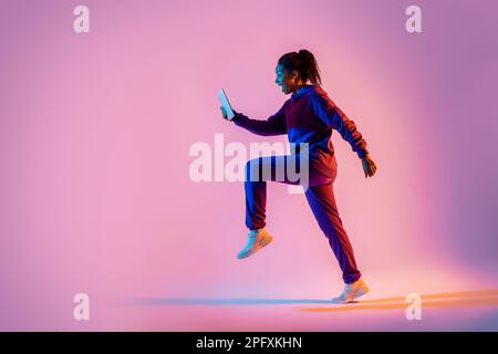 Prise de vue sur toute la longueur du corps d'une femme noire sautant ou courant, en utilisant un téléphone portable sur fond rose fluo, vue latérale, espace de copie Banque D'Images