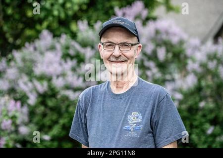 Beau portrait d'un homme âgé heureux qui aime les corvettes de Ford avec des lilas pourpres dans le bokeh en arrière-plan le soir de printemps. Banque D'Images