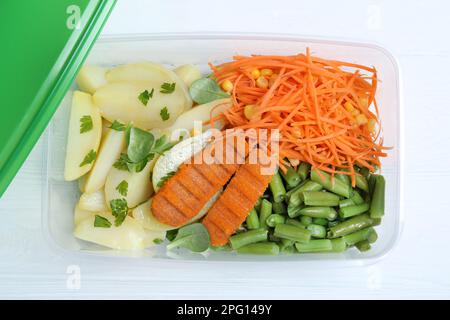 Pommes de terre savoureuses avec des côtelettes et des légumes dans un récipient en plastique sur une table en bois blanc, vue de dessus Banque D'Images
