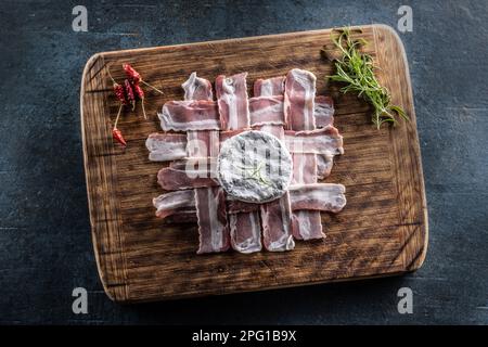 Vue de dessus d'une planche en bois sur une surface rustique sombre avec des tranches de prosciutto, du fromage camembert, de la brindille de romarin et des piments frais. Banque D'Images