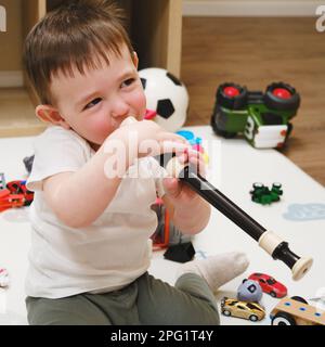 Bébé joue la flûte assise sur le sol dans la chambre des enfants. Enfant garçon jouant des instruments de musique. Enfant âgé d'un an et de huit mois Banque D'Images