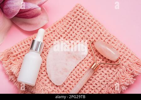 Pierre de Gua sha, rouleau de visage, produits cosmétiques et fleur de magnolia sur une serviette sur table rose. Vue de dessus, plan d'agencement. Banque D'Images