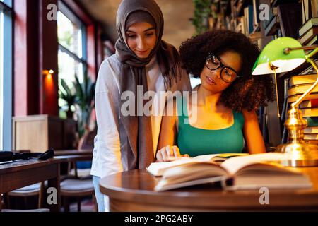 Deux adolescentes de différentes nationalités sont à la bibliothèque et lisent des livres. Fille américaine africaine avec des lunettes montre fille musulmane dans hijab à la page. Banque D'Images