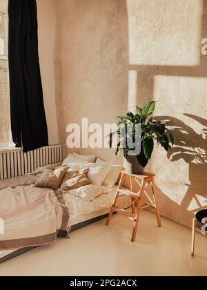 Chambre ensoleillée avec lit et grande fleur à côté. Literie claire et beaucoup d'oreillers. Le pot de plantes se trouve sur un élégant support en bois. Murs en plâtre beige et Banque D'Images