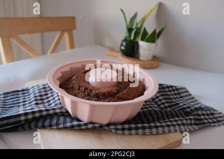 Gâteau cuit dans un moule en silicone, gâteau cuit avec cacao dans un moule en silicone rond rose sur un tissu de cuisine à carreaux de couleur foncée sur une table de cuisine blanche. Banque D'Images