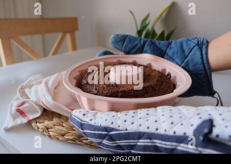 Gâteau au four, gâteau au four dans un moule en silicone rond rose entre deux mains sur un tissu blanc de cuisine et napperon dans la cuisine. La photo de concept. Banque D'Images