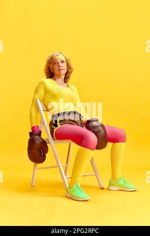 Femme sportive âgée en uniforme coloré assise sur une chaise en gants de boxe rétro, posant sur fond jaune de studio. Concept de sport et Banque D'Images