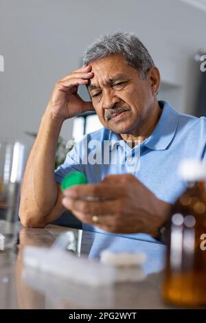 Gros plan d'un homme aîné triste, tête en main, regardant la bouteille prescrite sur la table à la maison Banque D'Images