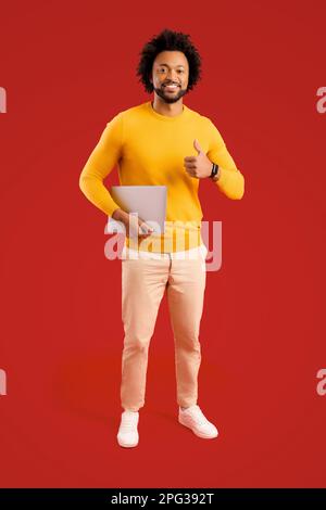 Homme souriant tenant un ordinateur portable dans les mains et homme indépendant souriant et charmant se tenant isolé sur fond rouge Banque D'Images