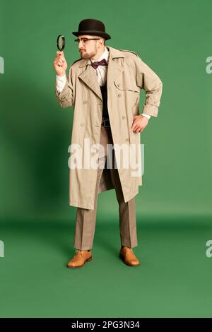 Portrait complet d'un jeune homme beau en trench-coat élégant et chapeau regardant dans la loupe sur fond vert de studio Banque D'Images