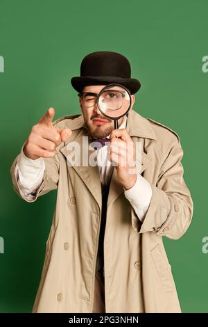 Jeune homme à l'image d'un détective, portant un trench-coat et un chapeau élégants, regardant dans la loupe sur fond vert de studio Banque D'Images