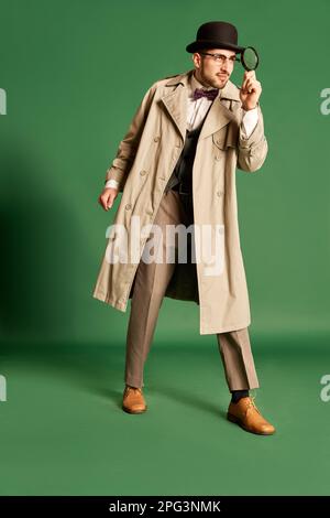 Portrait complet d'un jeune homme, détective dans un élégant trench coat et chapeau regardant dans la loupe sur fond vert studio Banque D'Images