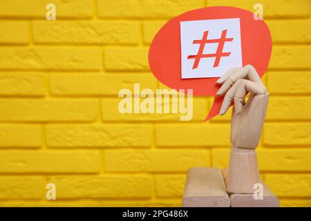 Main de mannequin en bois tenant une bulle de discours en papier avec un symbole de hashtag près du mur de brique jaune. Espace pour le texte Banque D'Images