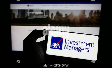 Personne détenant un téléphone portable avec logo de la société d'investissement AXA Investment Managers à l'écran en face de la page web business. Mise au point sur l'affichage du téléphone. Banque D'Images