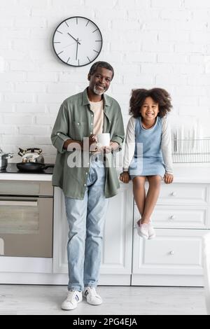 grand-père afro-américain d'âge moyen tenant une tasse de café près d'un enfant joyeux assis sur le plan de travail de la cuisine, image de stock Banque D'Images