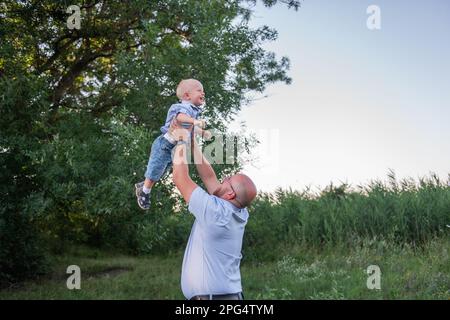 Homme chauve avec des lunettes jette l'enfant dans l'air du ciel. Père en jeans joue, embrassez son fils dans la nature en dehors de la ville. Un petit garçon rit, s'amuse bien Banque D'Images