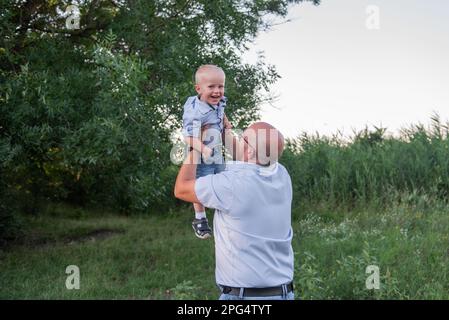 Homme chauve avec des lunettes jette l'enfant dans l'air du ciel. Père en jeans joue, embrassez son fils dans la nature en dehors de la ville. Un petit garçon rit, s'amuse bien Banque D'Images