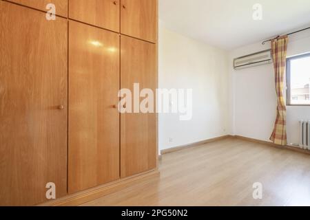 Cette chambre vide est dotée de parquet marron clair, d''une armoire intégrée avec des portes battantes en bois assorties, d''une fenêtre avec des rideaux et des murs blancs Banque D'Images