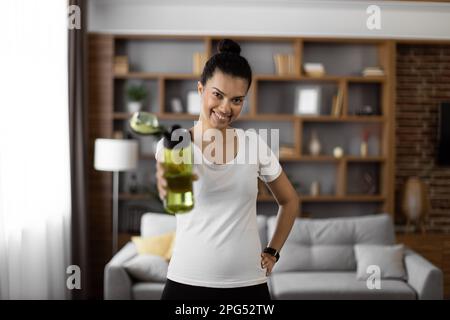 Une femme américaine positive tenant une bouteille d'eau de sport et essuyant la sueur du front pendant la pause entre les exercices à la maison. Jeune femme aux cheveux foncés Banque D'Images