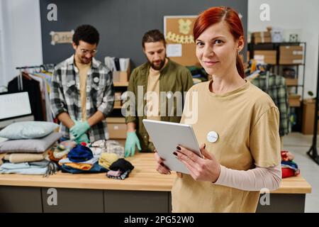 Femme mature leader de l'organisation de bénévolat avec une tablette contre de jeunes volontaires préparant des vêtements pour les personnes dans le besoin Banque D'Images