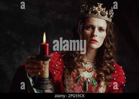 reine médiévale en robe rouge avec bougie et couronne sur fond gris foncé. Banque D'Images