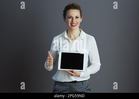 Souriante élégante petite entreprise propriétaire femme dans un chemisier blanc montrant tablette PC écran vide et pouces vers le haut sur fond gris. Banque D'Images