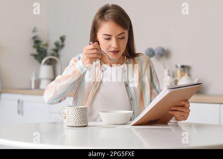 Belle femme avec un magazine en train de manger de la soupe à la table dans la cuisine Banque D'Images