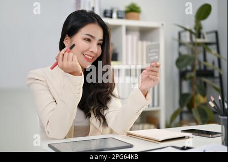 Charmante et belle employée de bureau asiatique du millénaire appliquant du maquillage à son bureau dans le bureau. Banque D'Images