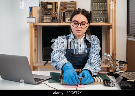 Le technicien répare la carte mère dans le centre de service. Femme réparateur travaillant avec un ordinateur avec une pièce en mains. Moniteurs et autres ordinateurs portables en arrière-plan Banque D'Images