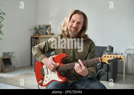 Portrait d'un guitariste mature avec de longs cheveux regardant l'appareil photo tout en jouant de la guitare en ligne Banque D'Images