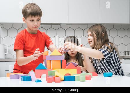 Des enfants positifs et concentrés de garçons et de deux filles jouant avec des blocs colorés et le château de construction à partir de figures géométriques sur table blanche. Créatif et Banque D'Images