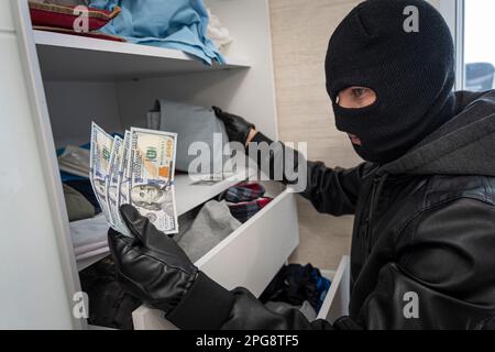 Un voleur dans un masque noir cherche un placard avec des vêtements à la recherche d'argent ou d'objets de valeur. Vol d'appartement. Une recherche de la maison. Petite criminalité Banque D'Images