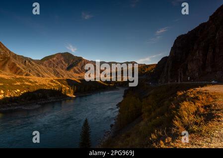 La route de montagne Chuisky Trakt va sous un rocher à l'ombre à côté de la rivière Katun en Altaï dans la soirée en Russie. Banque D'Images