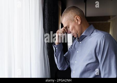 Portrait d'un homme qui se tient près de la fenêtre et tient sa tête d'une main, a un mal de tête ou est réfléchi. Banque D'Images