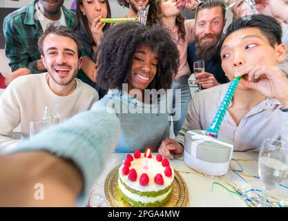 Joyeux groupe d'amis multiethniques prenant le selfie tout en ayant la fête d'anniversaire à la maison, en utilisant le téléphone mobile et en buvant du champagne Banque D'Images