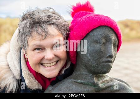 femme d'âge moyen souriant avec sculpture en métal de la personne avec chapeau de galet rose sur South Shields, Tyne et Wear UK Banque D'Images