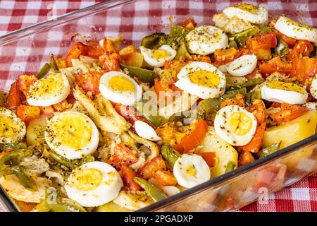 Gomes de sa morue rôtie dans de l'huile d'olive avec tomates, poivrons, oignons, œufs durs et origan. Dans un plat de cuisson rectangulaire en verre sur un damiré à Banque D'Images