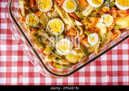 Gomes de sa morue rôtie dans de l'huile d'olive avec tomates, poivrons, oignons, œufs durs et origan. Dans un plat de cuisson rectangulaire en verre sur un damiré à Banque D'Images