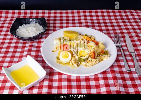 Gomes de sa morue rôtie dans de l'huile d'olive avec tomates, poivrons, oignons, œufs durs et origan. Dans un plat en porcelaine blanche sur une nappe à carreaux Banque D'Images