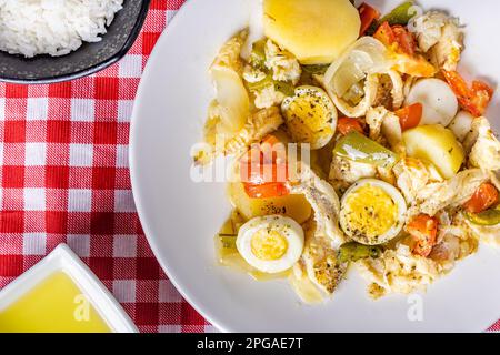 Gomes de sa morue rôtie dans de l'huile d'olive avec tomates, poivrons, oignons, œufs durs et origan. Dans un plat en porcelaine blanche sur une nappe à carreaux Banque D'Images