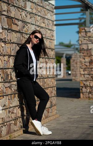 Une fille aux cheveux longs dans un survêtement noir et des lunettes de soleil. Un modèle dans une tenue de sport. Une belle fille pose sportily Banque D'Images