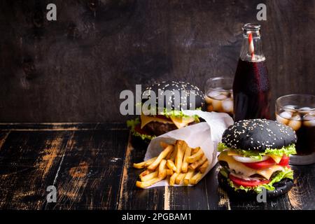 Ensemble de hamburgers noirs avec patty de viande, fromage, tomates, mayonnaise, frites et verre de soda au cola froid avec glace. Fond rustique en bois sombre. Banque D'Images