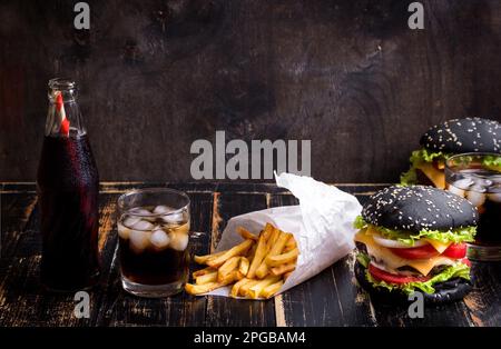 Ensemble de hamburgers noirs avec patty de viande, fromage, tomates, mayonnaise, frites et verre de soda au cola froid avec glace. Fond rustique en bois sombre. Banque D'Images