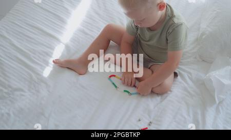 Vue de haut en bas d'un enfant caucasien recueille les formes géométriques d'un constructeur magnétique tout en étant assis à la maison sur un lit. Le garçon combine un jeu de loisirs Banque D'Images