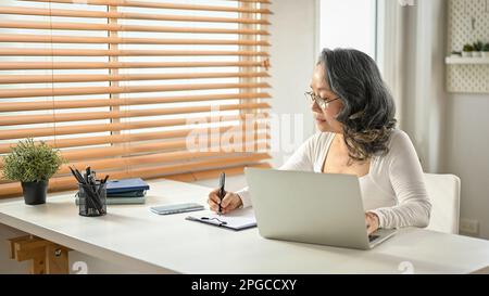 Professionnelle et concentrée 60s femme d'affaires asiatique dans des vêtements décontractés travaillant sur ses tâches d'affaires dans son bureau. concept d'homme d'affaires Banque D'Images