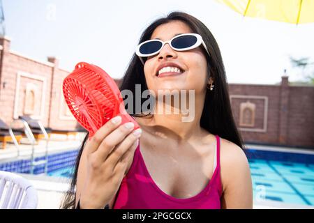 Jeune adolescente indienne portant des lunettes de soleil appréciant le flux d'air du ventilateur électrique portable et refroidissant son corps extérieur dans la journée ensoleillée. Chaleur estivale. Banque D'Images