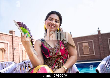 Belle jeune femme indienne utilisant un ventilateur main ondulée de papier assis à l'extérieur dans la journée ensoleillée souffrant de temps chaud d'été ou de haute température, Banque D'Images