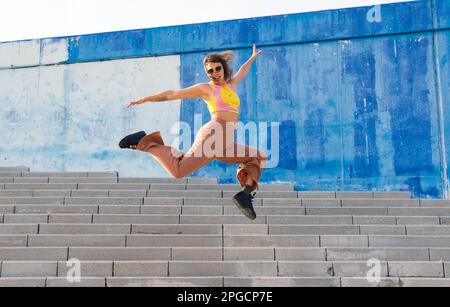 Active jeune femme dans des vêtements décontractés et des lunettes de soleil sautant au-dessus des escaliers avec les bras étirés tout en regardant l'appareil photo et en souriant dans la rue Banque D'Images